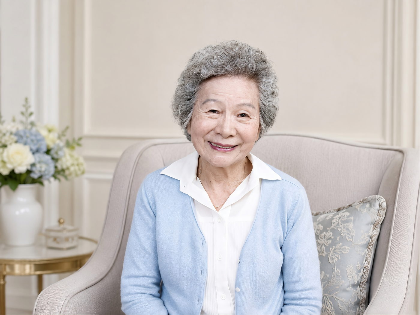 An older East Asian woman in a pale blue cardigan and white blouse sits in an upholstered armchair in a bright, calm room.