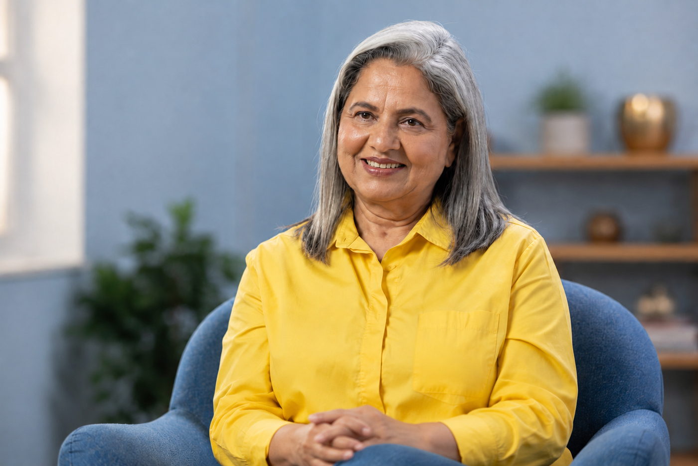 An older South Asian woman with long grey hair, wearing a yellow shirt, sits in a soft blue armchair at home.